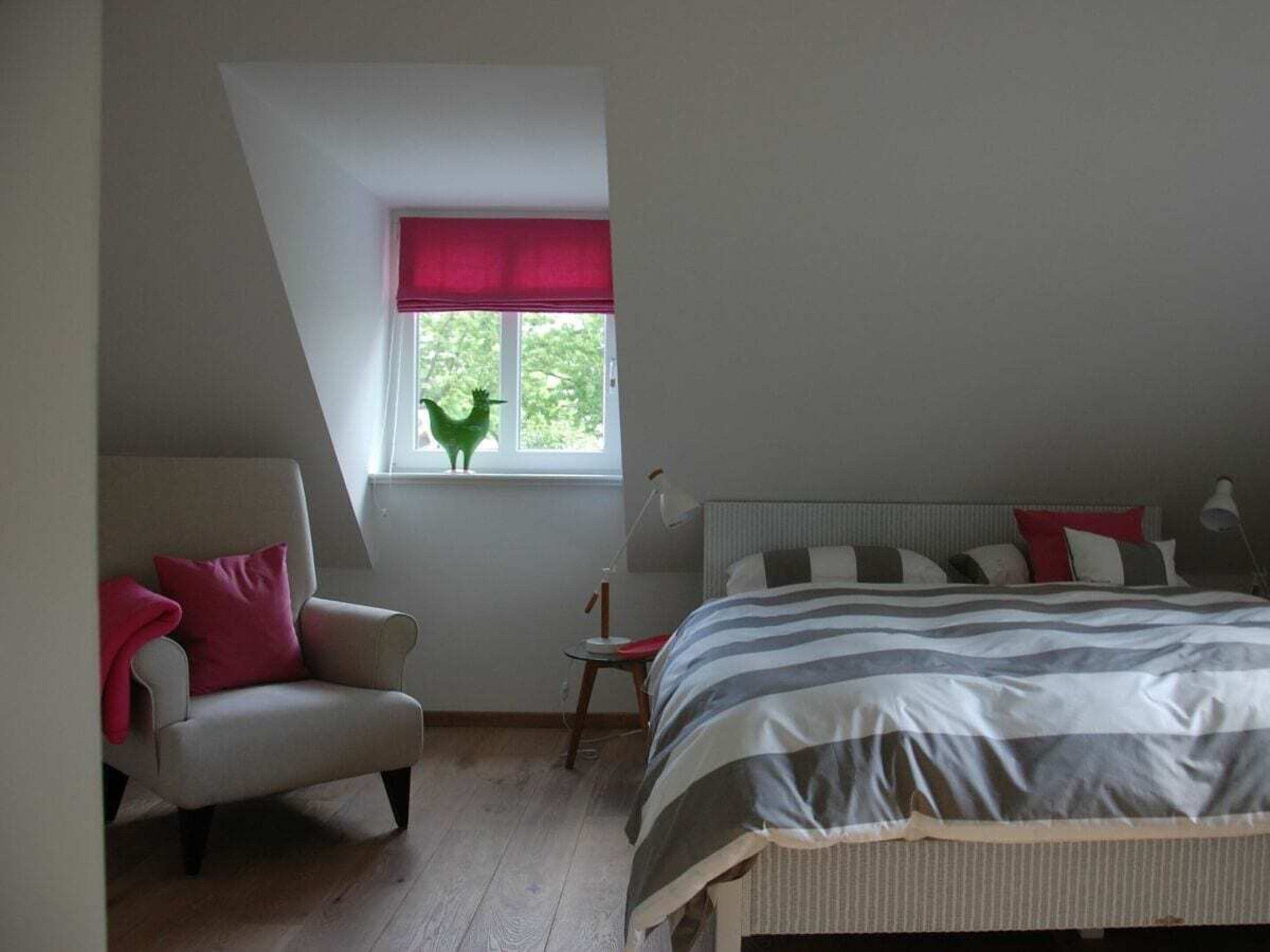 A bedroom with a sloped ceiling, a bed with striped bedding, a chair with a red pillow, a small table with a lamp, and a window with a red blind.