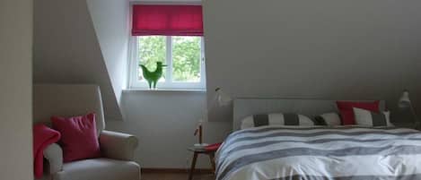A bedroom with a sloped ceiling, a bed with striped bedding, a chair with a red pillow, a small table with a lamp, and a window with a red blind.