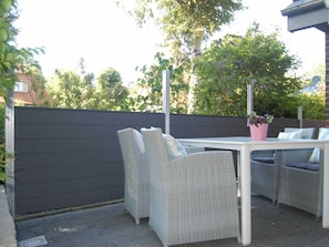 Une terrasse extérieure avec une table et des chaises en osier, entourée d’une clôture sombre et de verdure.