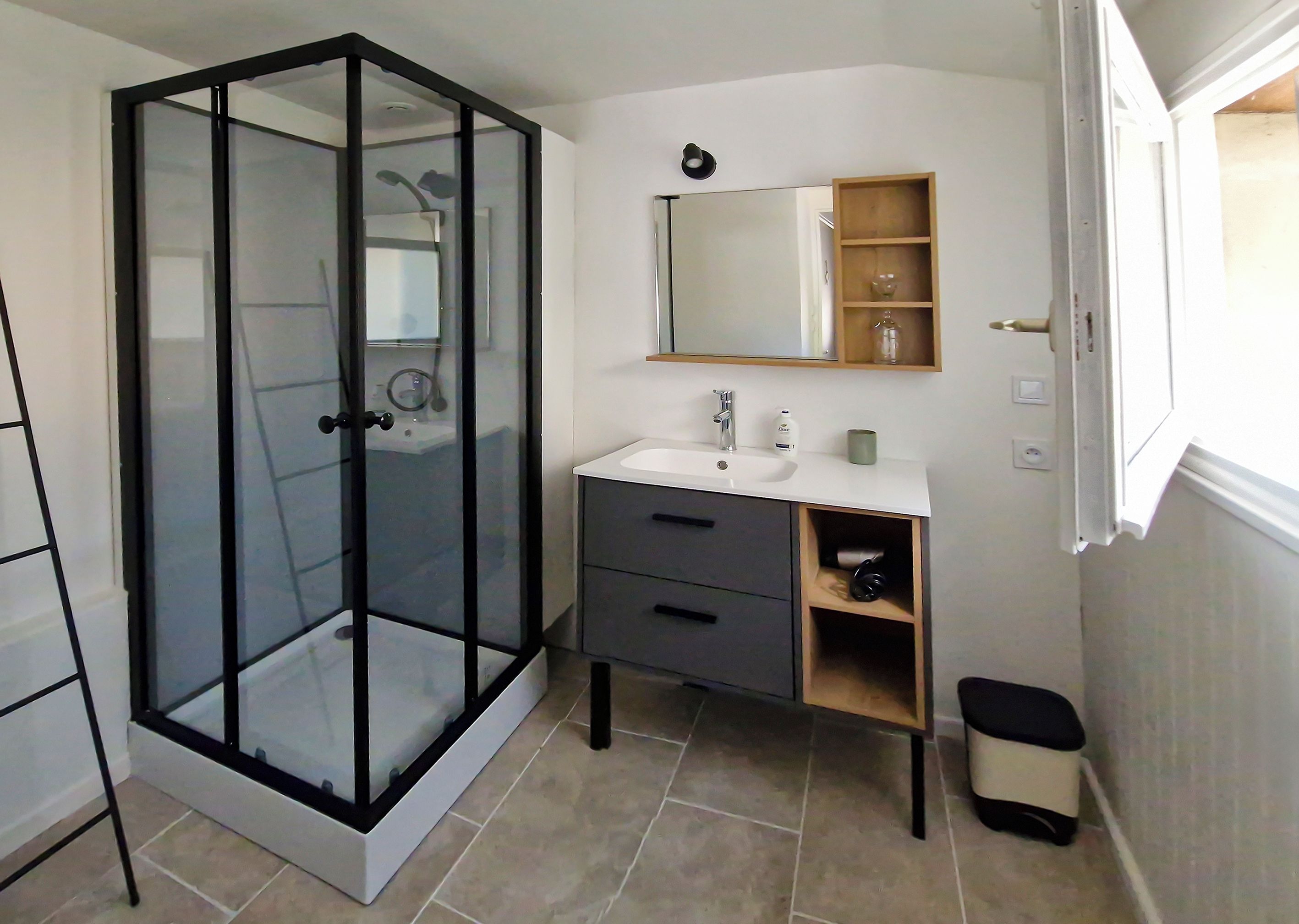 Une salle de bain avec une douche à parois de verre, un lavabo blanc, un miroir et une petite étagère contenant des articles de toilette.