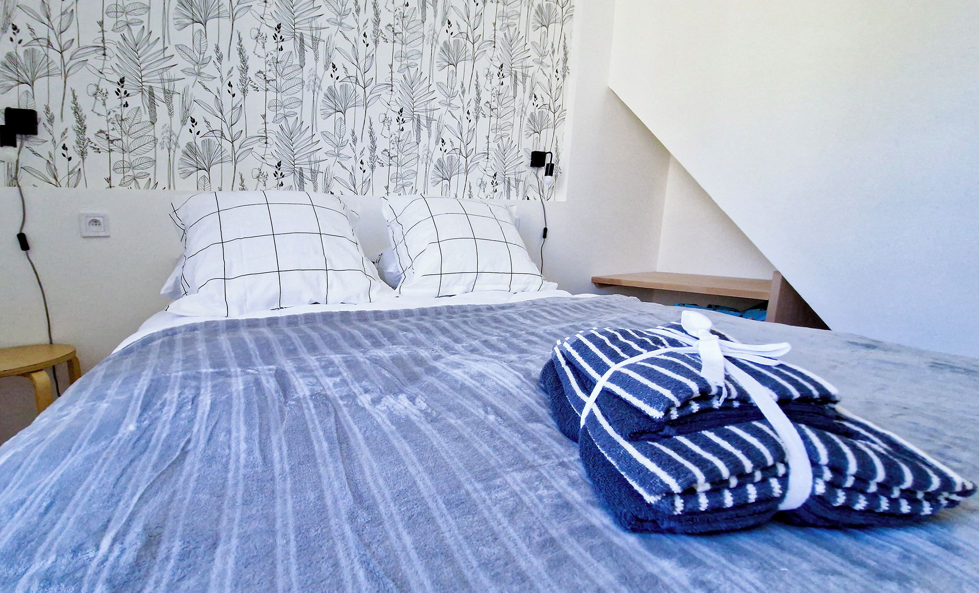 Un lit bien fait, recouvert d’une couverture à rayures bleues, avec un oreiller à motifs géométriques et une serviette pliée posée sur le lit.