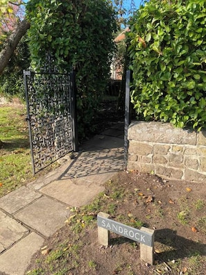 A gated entrance with a metal gate, a stone wall, and a sign indicating the name 'Sandrock.' - Cosy Country Cottage with Log Burner & Garden Near Petworth (Northchapel)