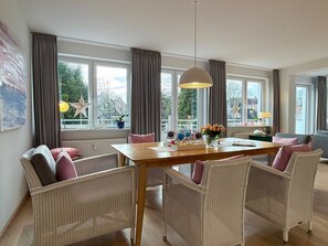 A dining area with a wooden table and wicker chairs, large windows with curtains, and a view of trees outside. - Above the Rooftops of Wyk/ Near the Beach (Wyk auf Foehr)