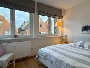 A bedroom with a bed, a chair, a window with a view of buildings, and a lamp. - Above the Rooftops of Wyk/ Near the Beach (Wyk auf Foehr)