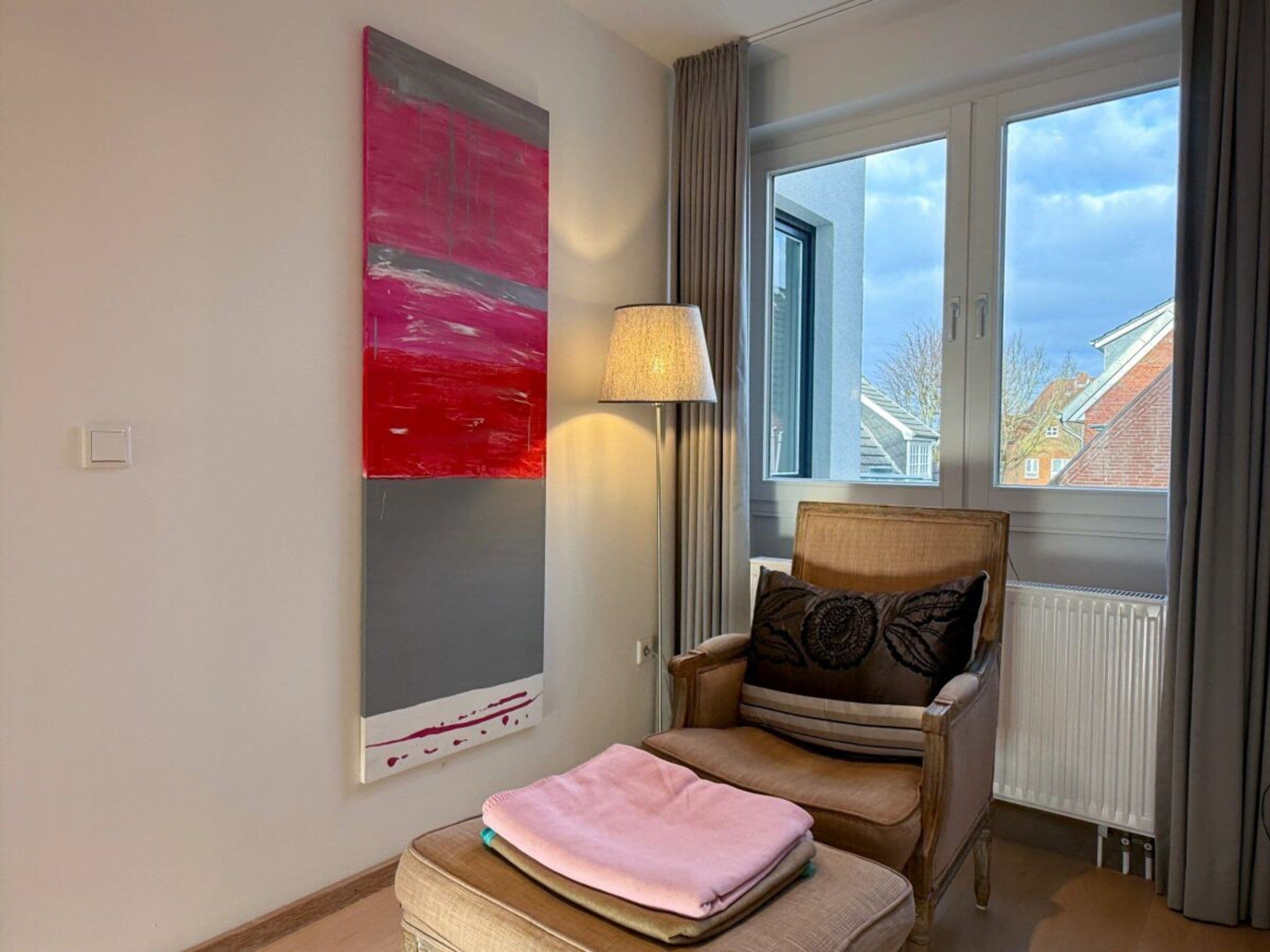Een kamer met een groot abstract schilderij, een stoel met een kussen, een nachtkastje met een roze object, en een raam met uitzicht op gebouwen.