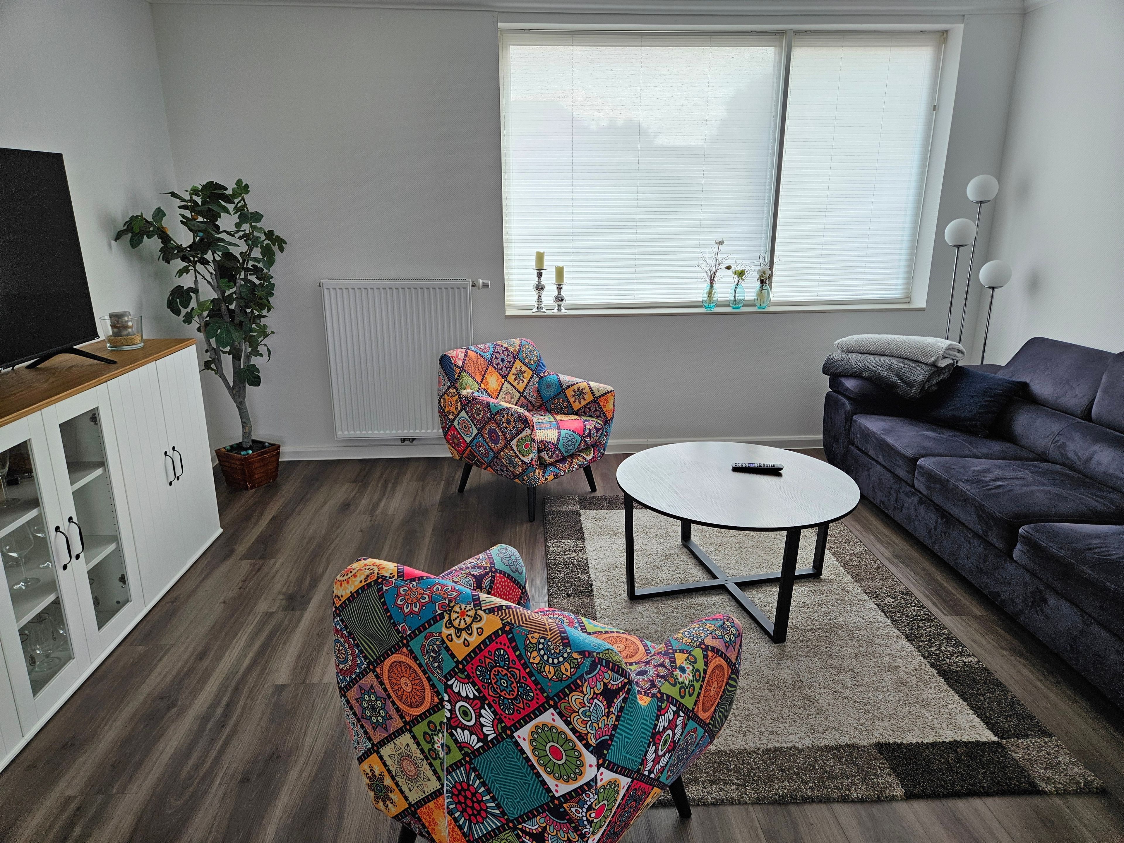 Una sala de estar con un sofá, dos sillones, una mesa de centro, un televisor sobre un mueble, una planta en maceta y una ventana con persianas.