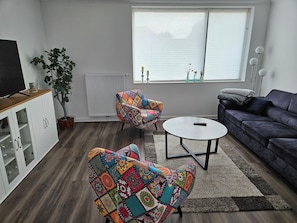 Ein Wohnzimmer mit einer Couch, zwei Sesseln, einem Couchtisch, einem Fernseher auf einem Schrank, einer Zimmerpflanze und einem Fenster mit Jalousien.