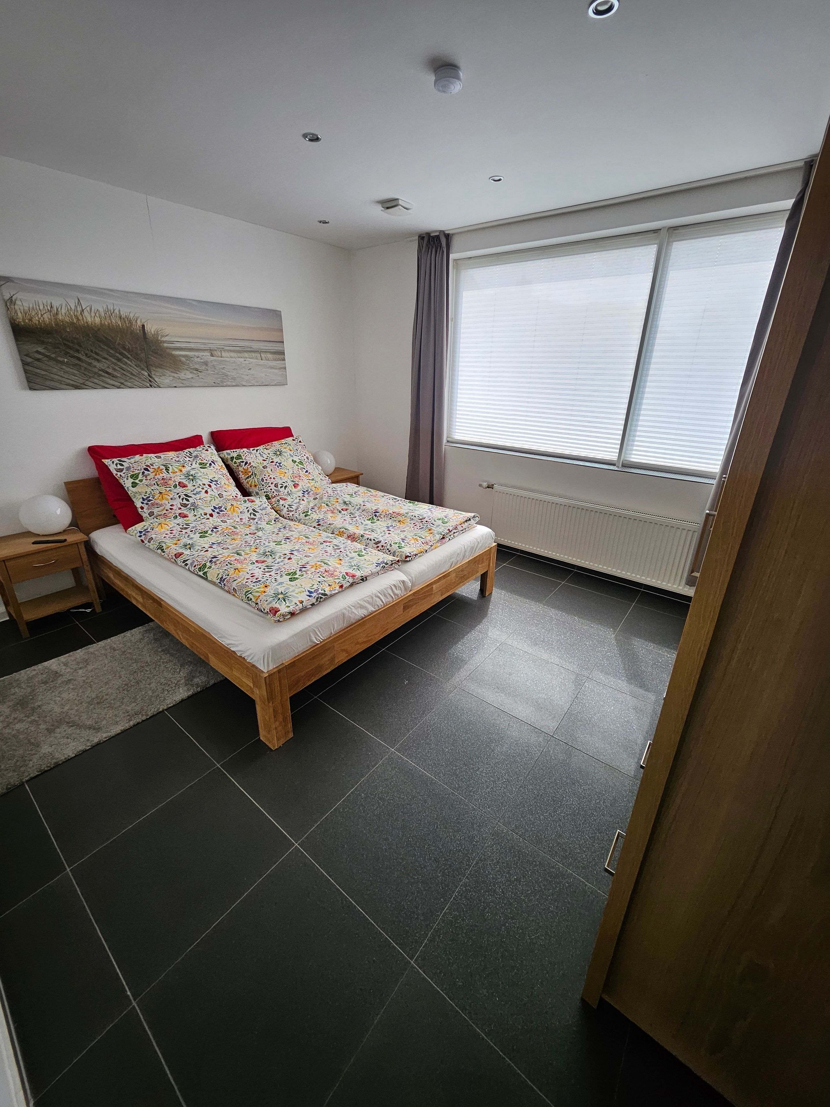 Een slaapkamer met een bed, nachtkastje, lamp, radiator en een raam met jaloezieën.
