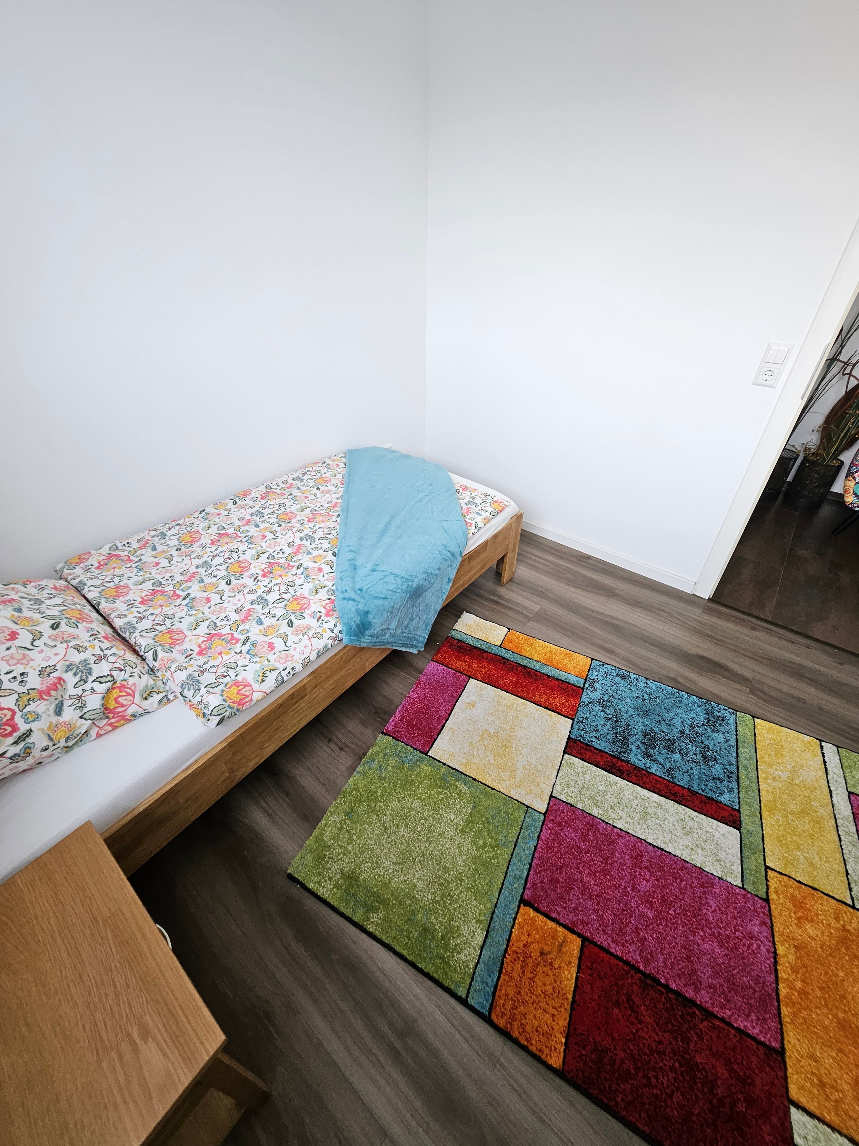 Pieni huone, jossa on yksi sänky, puulattia, värikäs matto ja kukkakuviollinen päiväpeite.