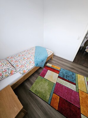 A small, single-bed room with a wooden floor, a colorful rug, and a floral-patterned bedspread.