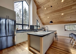 A modern kitchen with a central island, wooden ceiling, and large windows. - La Tanière de l'Ours BJÖRN HÎ, Refuge & Réconfort (Saint-Côme)