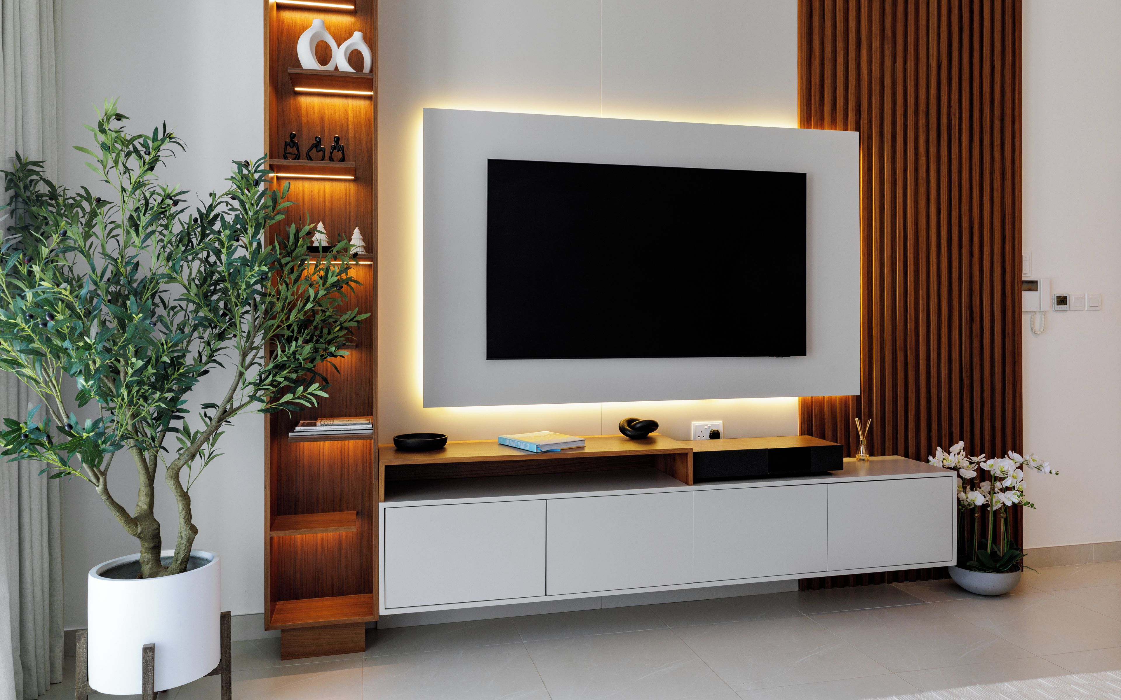 A modern living room with a large flat-screen TV mounted on a white cabinet, a potted plant, and a decorative shelf with figurines.