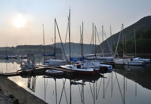 Una marina con varios veleros amarrados, agua tranquila y un paisaje montañoso al fondo.
