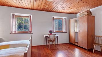 A room with a wooden ceiling, two windows, a bed, a table with a chair, and a wardrobe.
