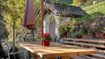 Un tavolo di legno con un vaso rosso, circondato da panche di legno, in un giardino con una casa rustica e vegetazione sullo sfondo.