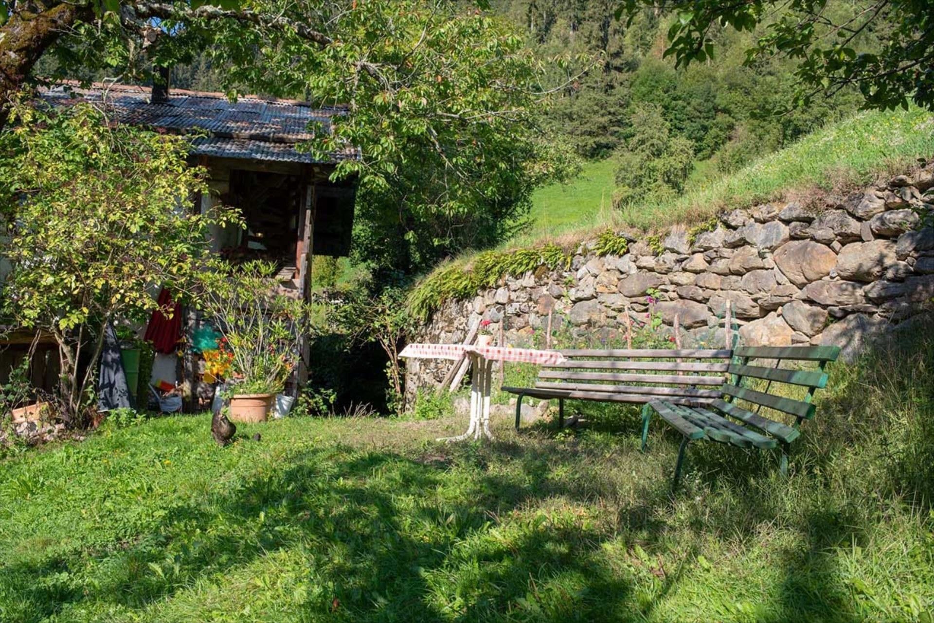 En rustik träbänk placerad vid en stenmur, omgiven av frodig grönska och ett litet hus med ett tak belagt med tegelpannor.