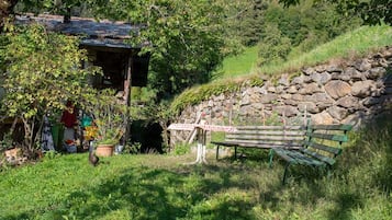 En rustik träbänk placerad vid en stenmur, omgiven av frodig grönska och ett litet hus med ett tak belagt med tegelpannor.