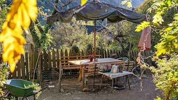 Ein Gartenbereich im Freien mit einem Holztisch und Bänken, umgeben von Bäumen und einem Blick auf die Berge.