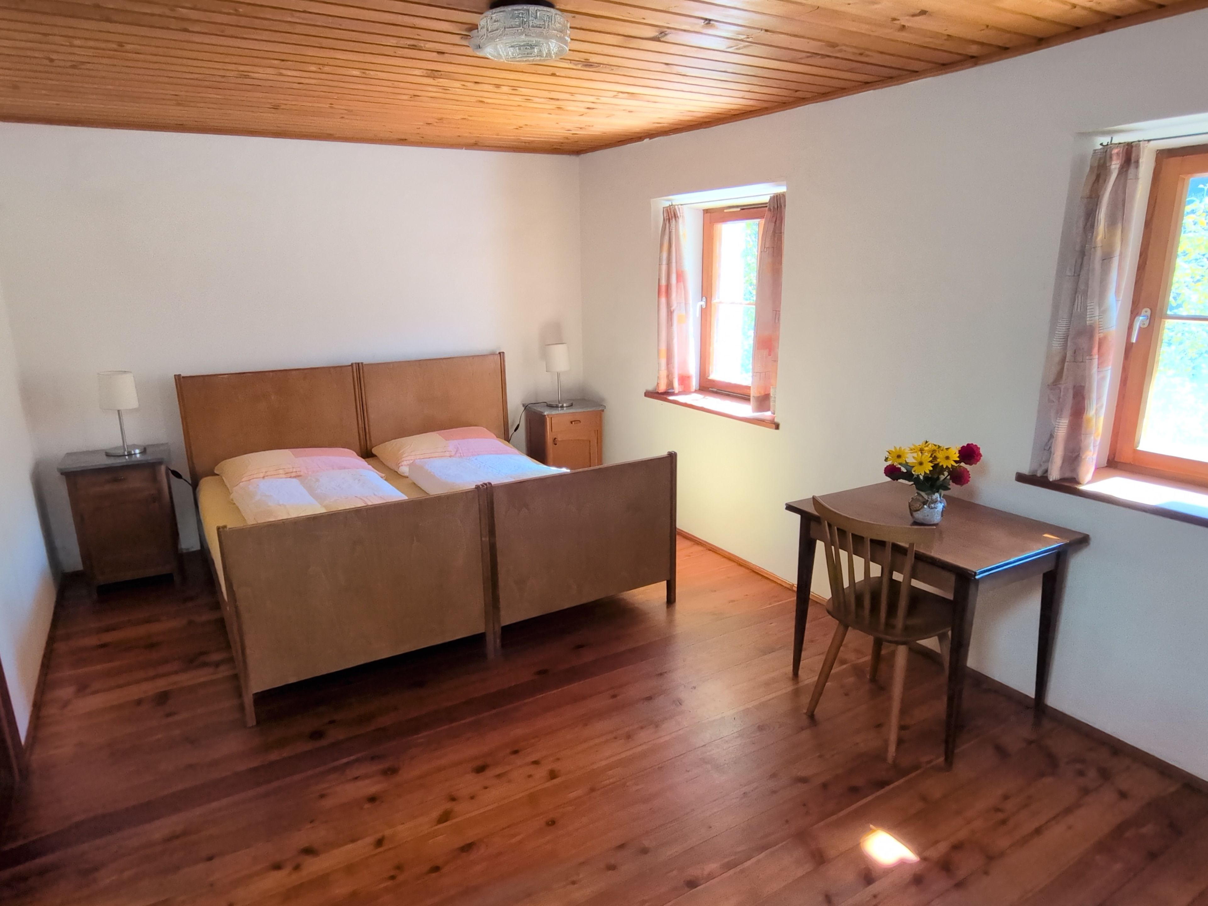 Ein Schlafzimmer mit Doppelbett, Nachttischen, einem Schreibtisch und einem Sessel. Es gibt zwei Fenster mit Vorhängen.