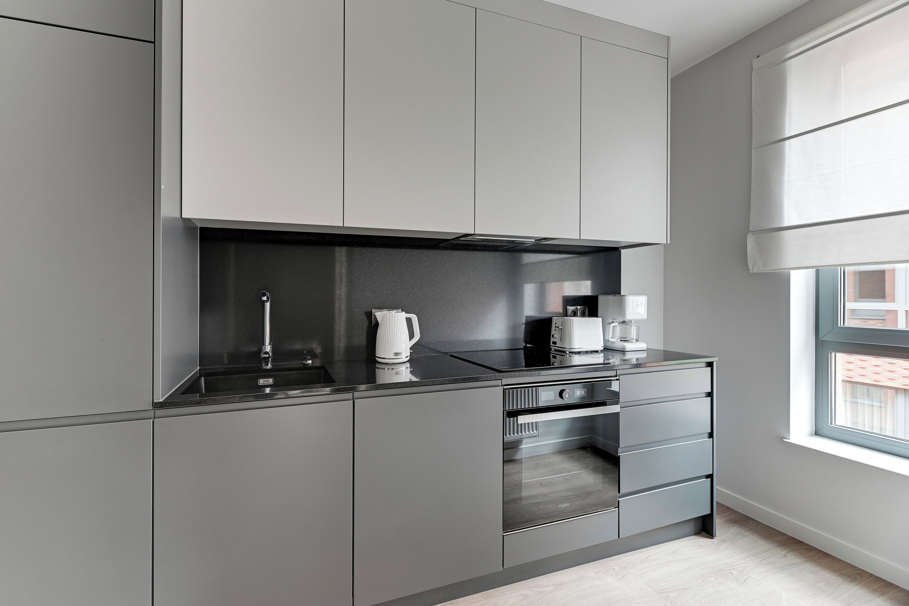 A modern kitchen with grey cabinetry, a built-in oven, and a window with blinds.