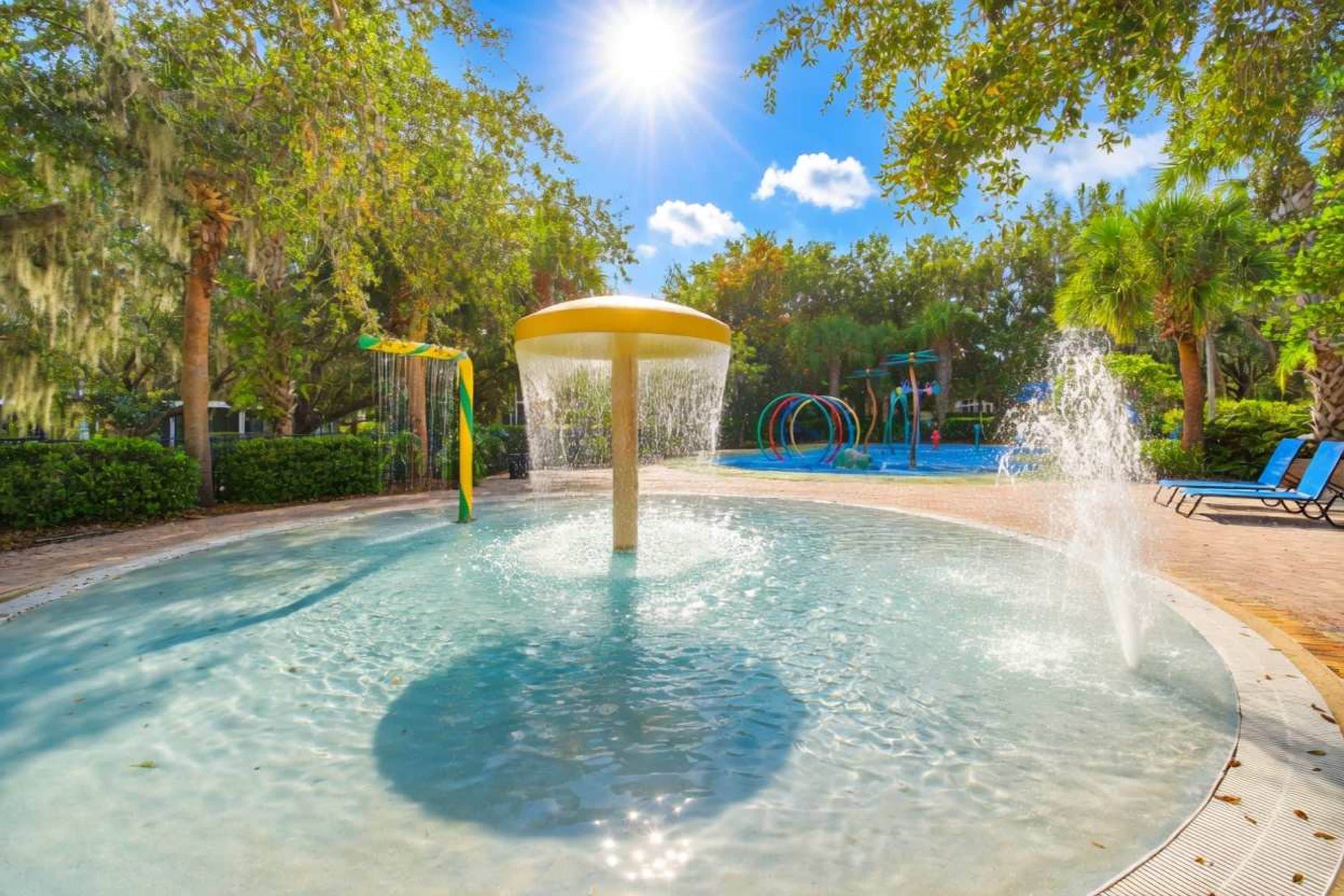 A water feature with a central fountain, surrounded by a paved area with lounge chairs and a playground.