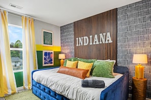 A bedroom with a bed, pillows, a wall with the word 'INDIANA', and a window with curtains. - Indy Escape Sauna Hot Tub Firepit Games (South Bend)