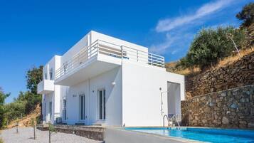 A modern white villa with a swimming pool, a stone wall, and a clear blue sky.