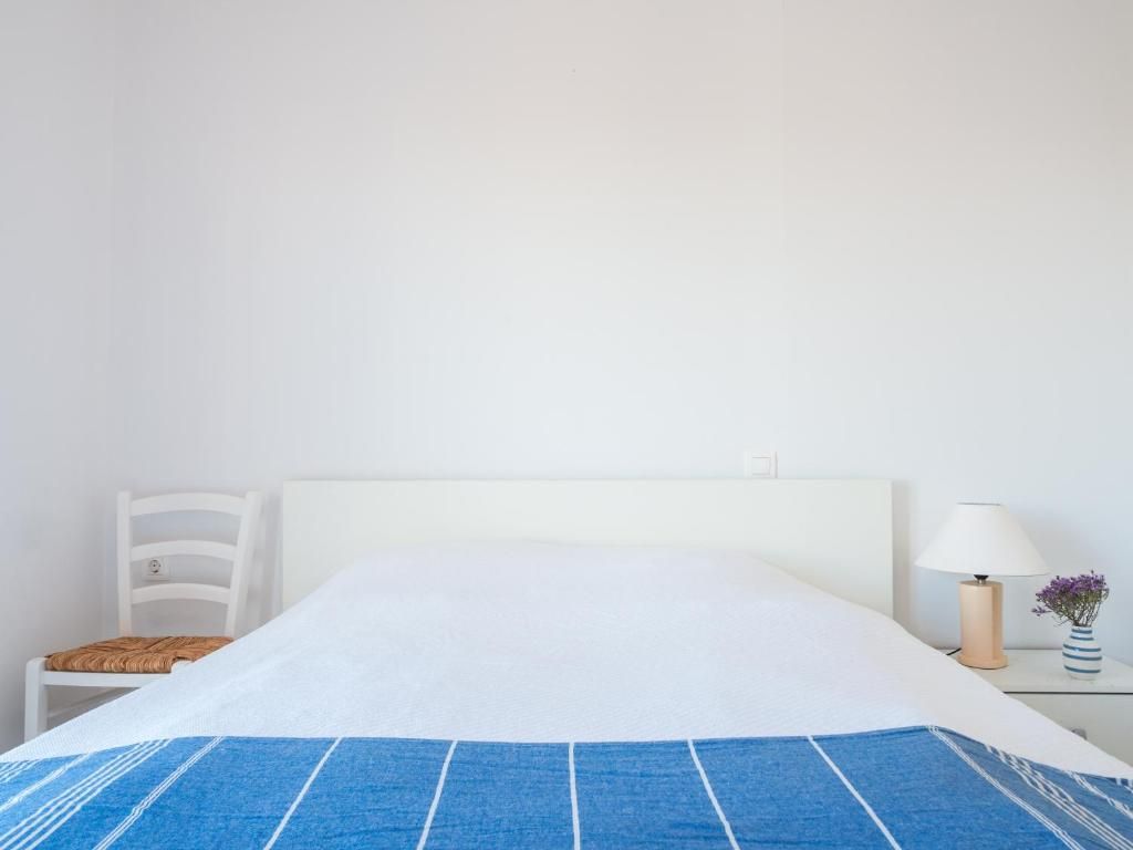 Una cama con cabecera blanca, una colcha a rayas azul y blanco, una silla blanca, una mesita de noche con una lámpara y un florero con flores.