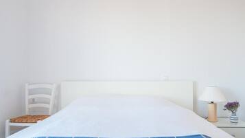 Una cama con cabecera blanca, una colcha a rayas azul y blanco, una silla blanca, una mesita de noche con una lámpara y un florero con flores.