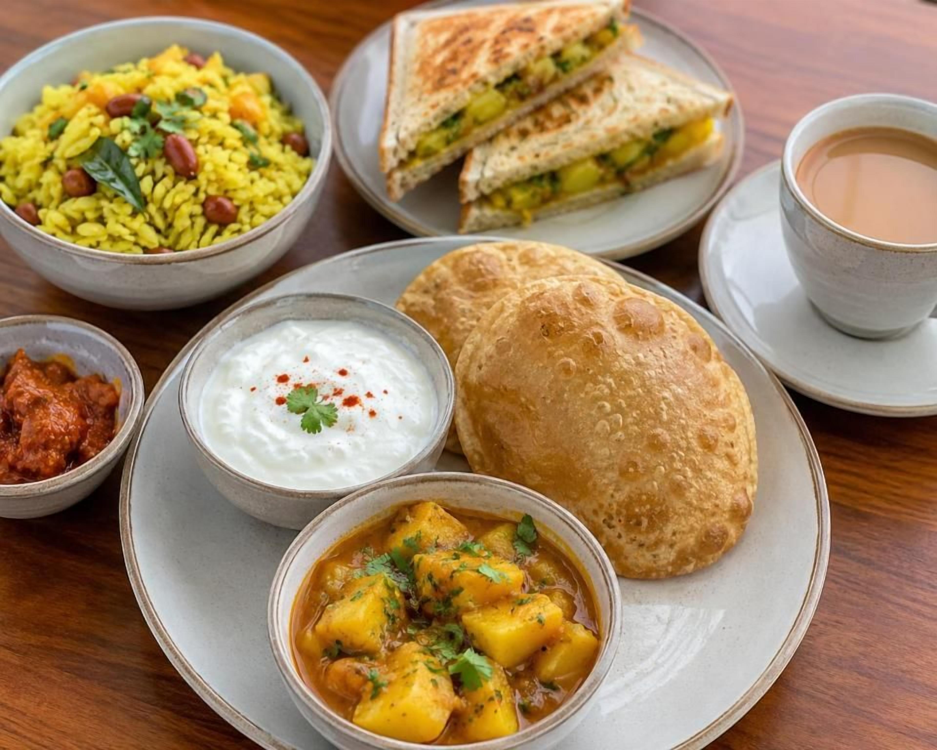 A table with a variety of dishes including rice, a sandwich, a flatbread, a bowl of curry, a bowl of yogurt, and a cup of tea.