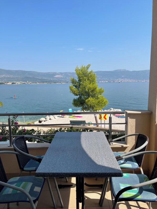 A balcony with a table and chairs, overlooking a beach and a body of water with a distant mountain range.