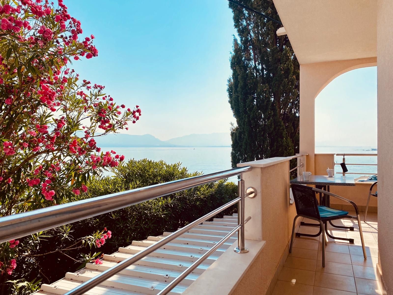 A balcony with a metal railing, a table with chairs, and a view of the sea and mountains.