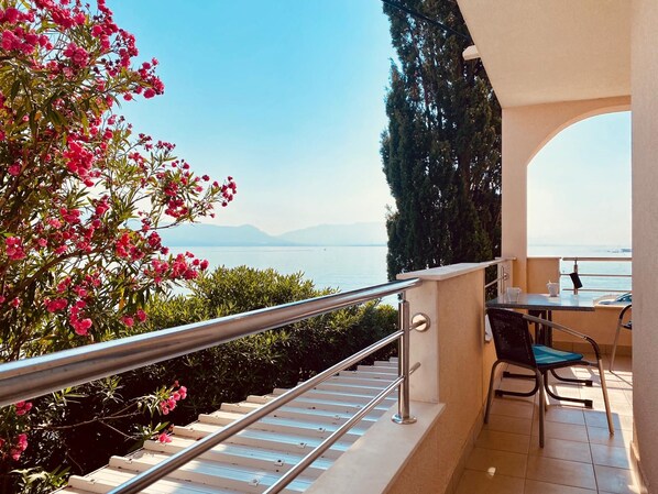 A balcony with a metal railing, a table with chairs, and a view of the sea and mountains.