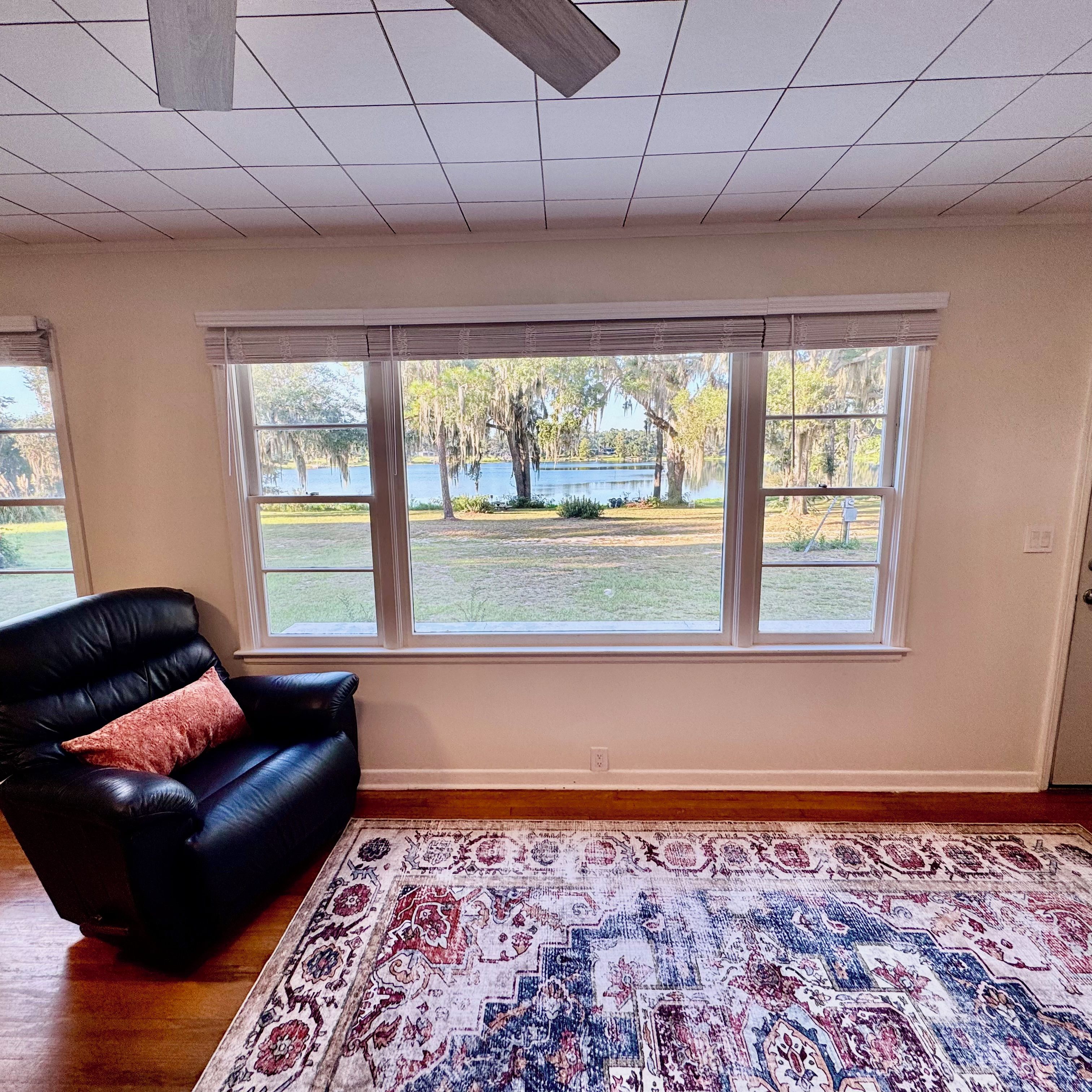 A room with a black leather sofa, a patterned rug, and large windows offering a view of a lake and trees.