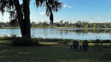Blick auf einen See mit zwei Adirondack-Stühlen, die dem Wasser zugewandt sind, umgeben von Gras und Bäumen.