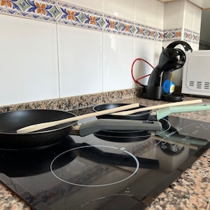 A kitchen with a stove, two frying pans, a wooden spatula, and a coffee maker. - Casa Resort Mi Alcazar by Rental Hero (Alcazares, Los)