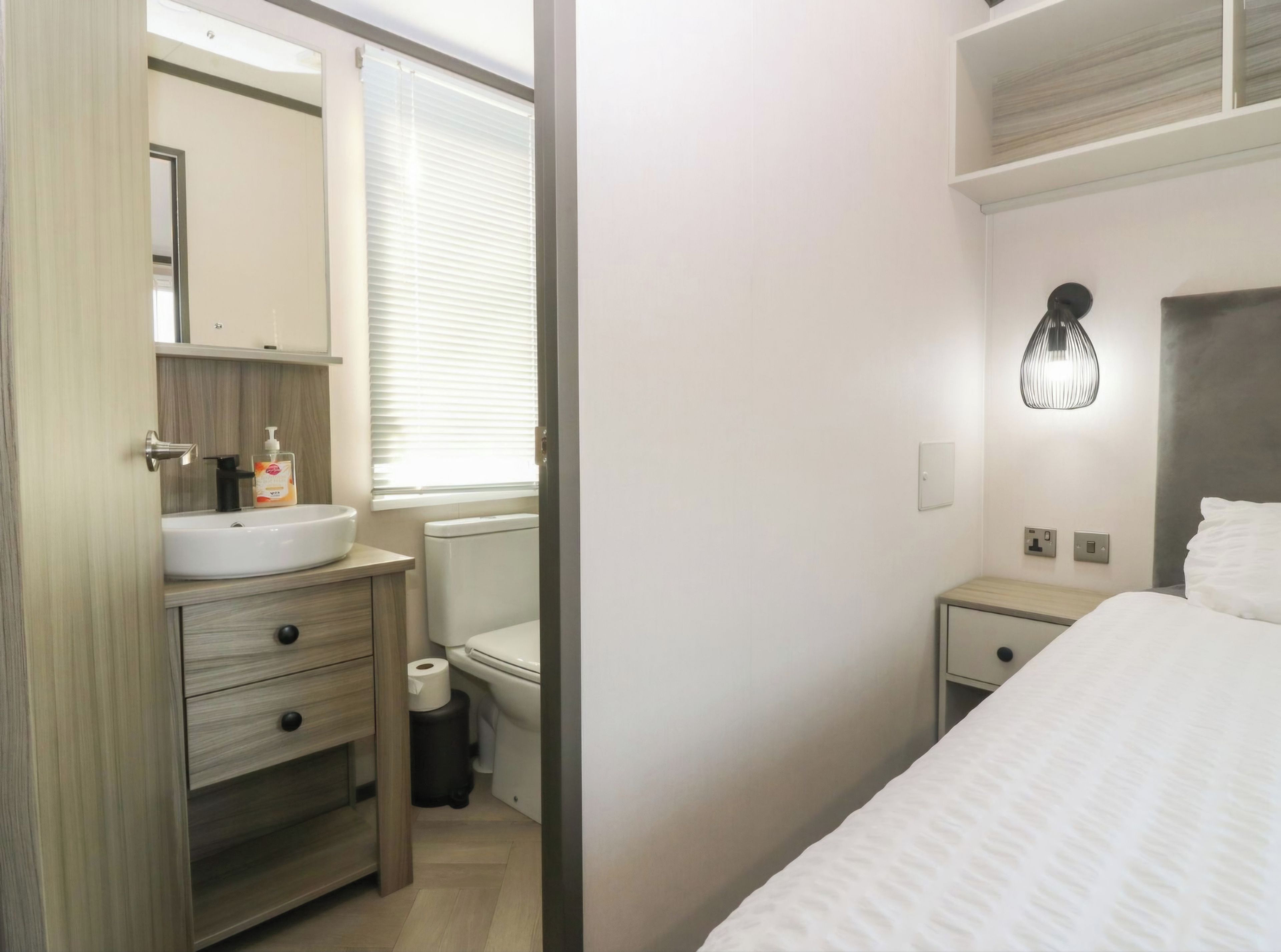 A hotel room with a bed, a bedside table, a nightstand, a bathroom with a sink, toilet, and a mirror.