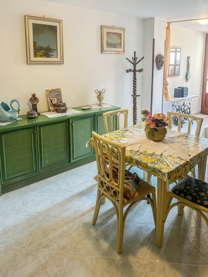 Een eetgedeelte met een groen kastje, rieten stoelen, een tafelkleed met bloemenmotief en kunst aan de muren.