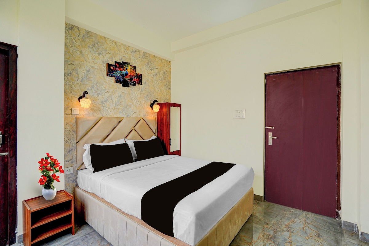 Habitación de hotel con cama, mesita de noche con un florero y una puerta, además de una pared decorada con un cuadro.