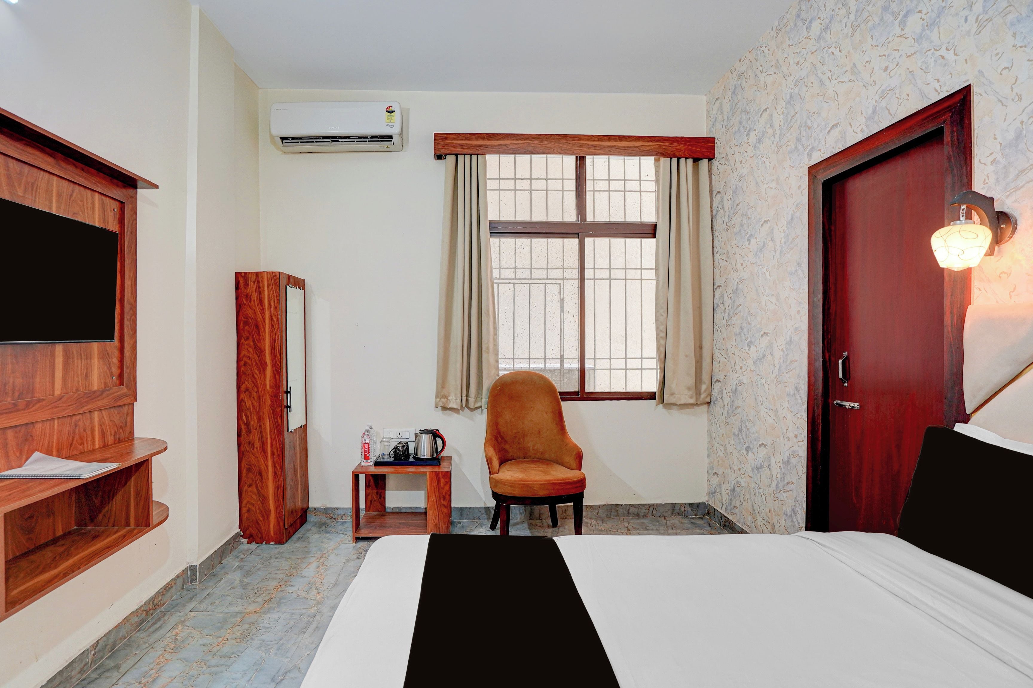 Quarto de hotel com cama, guarda-roupa de madeira, televisão, janela com cortinas, cadeira, mesa pequena com chaleira e garrafa, e porta com luminária.