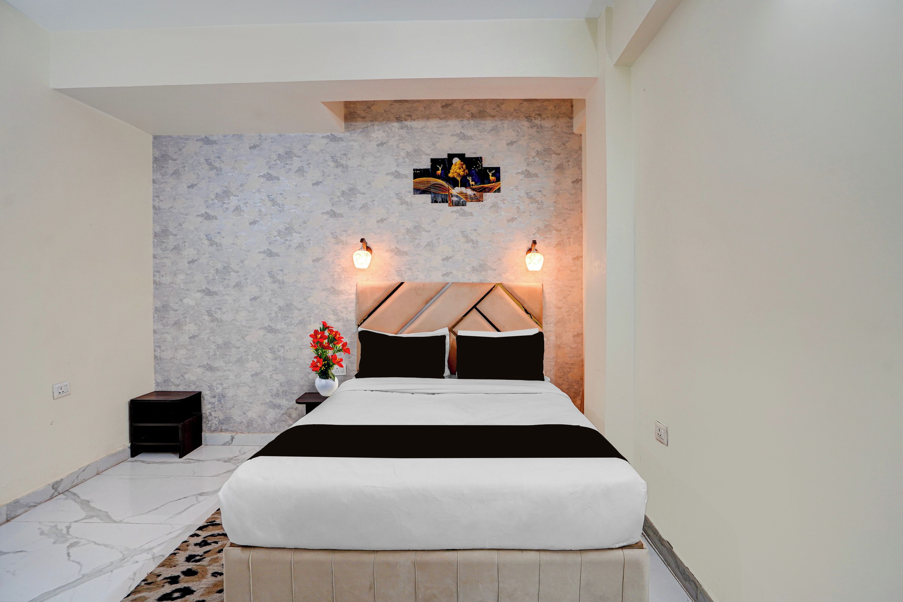Un dormitorio con cama, mesita de noche, un jarrón con flores y una lámpara de pared.