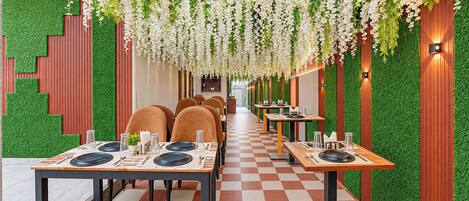 Un restaurant avec un sol à carreaux, des murs verts et des fleurs blanches suspendues. Les tables sont dressées avec des assiettes, des verres et des couverts.