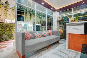 A modern interior with a grey sofa, a wooden reception desk, and large windows with greenery. - Collection O RPS More (Danapur)