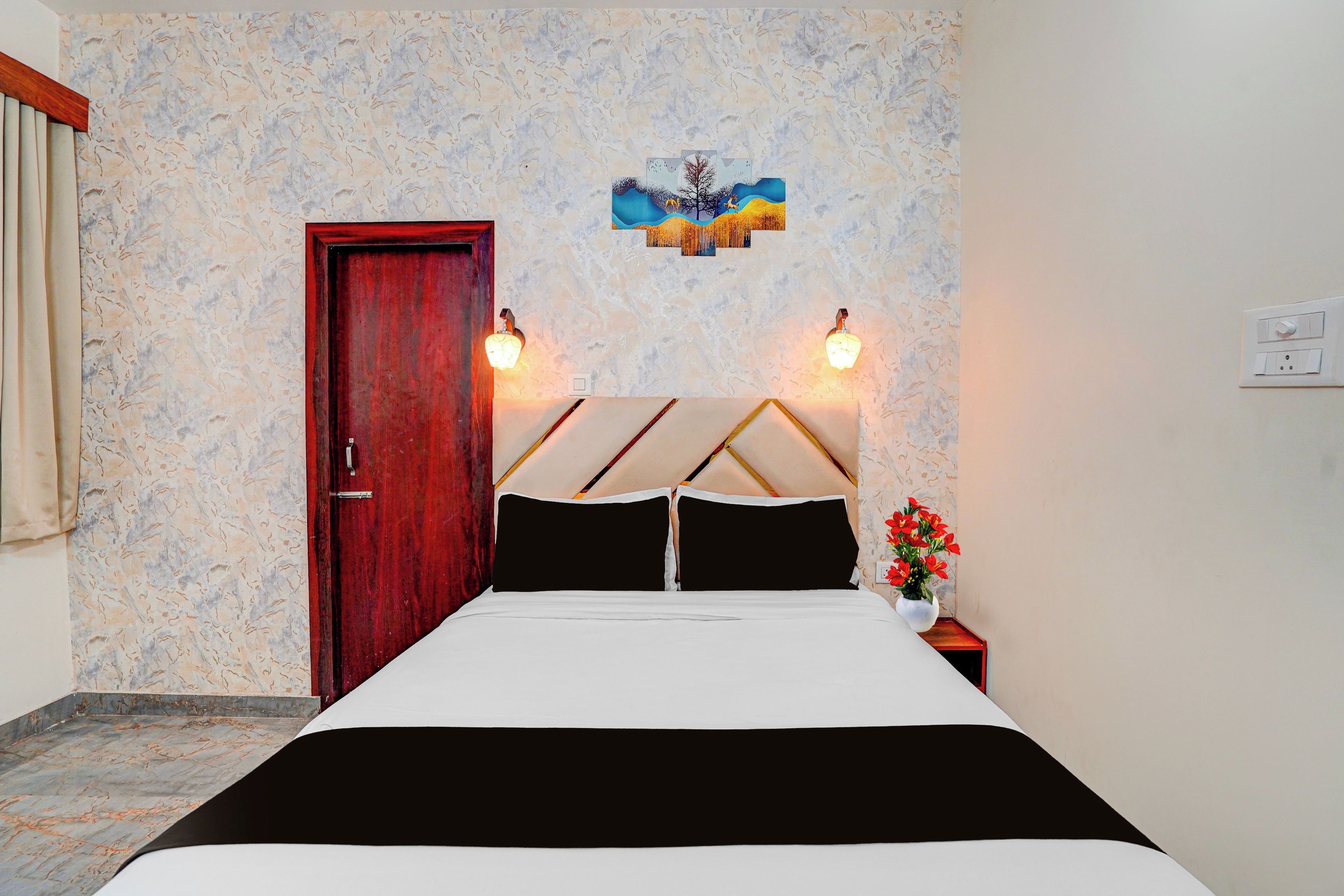 A bedroom with a bed, a wooden door, a wall-mounted light fixture, and a small table with a flower vase.