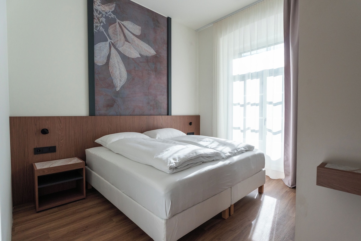A neatly made bed with white linens in a room with wooden flooring and a large wall-mounted artwork.