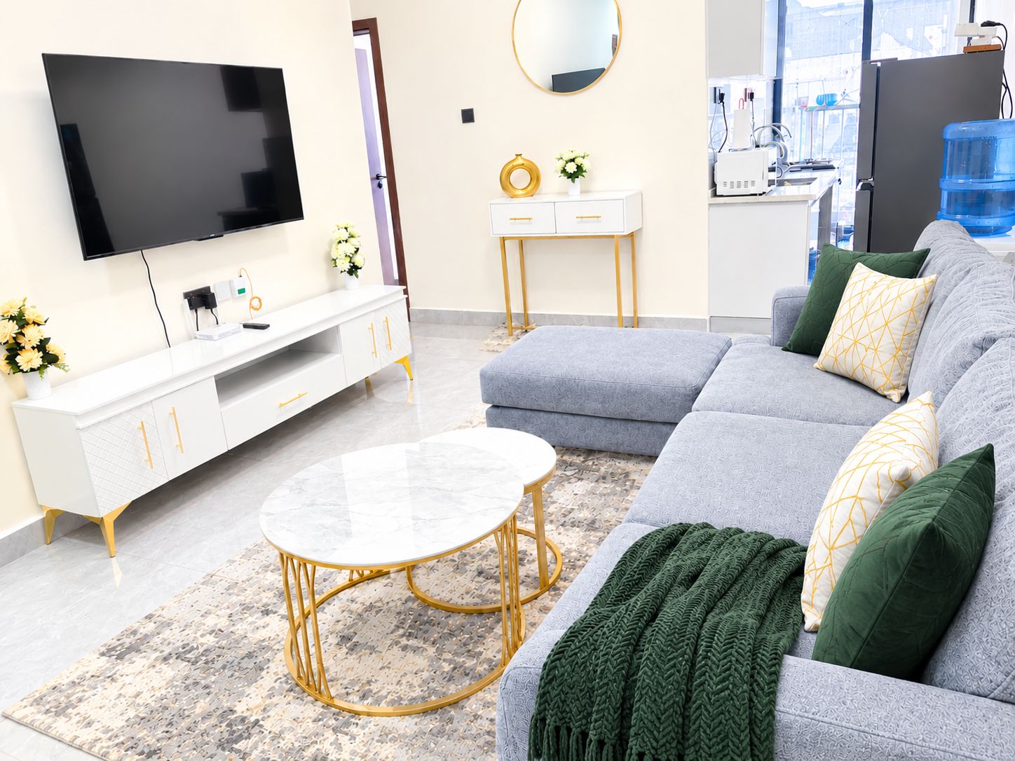 A modern living room with a flat-screen TV, a white console table, a grey sofa with green and yellow cushions, and a round coffee table with a gold base.
