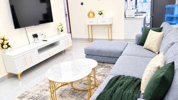 A modern living room with a flat-screen TV, a white console table, a grey sofa with green and yellow cushions, and a round coffee table with a gold base.