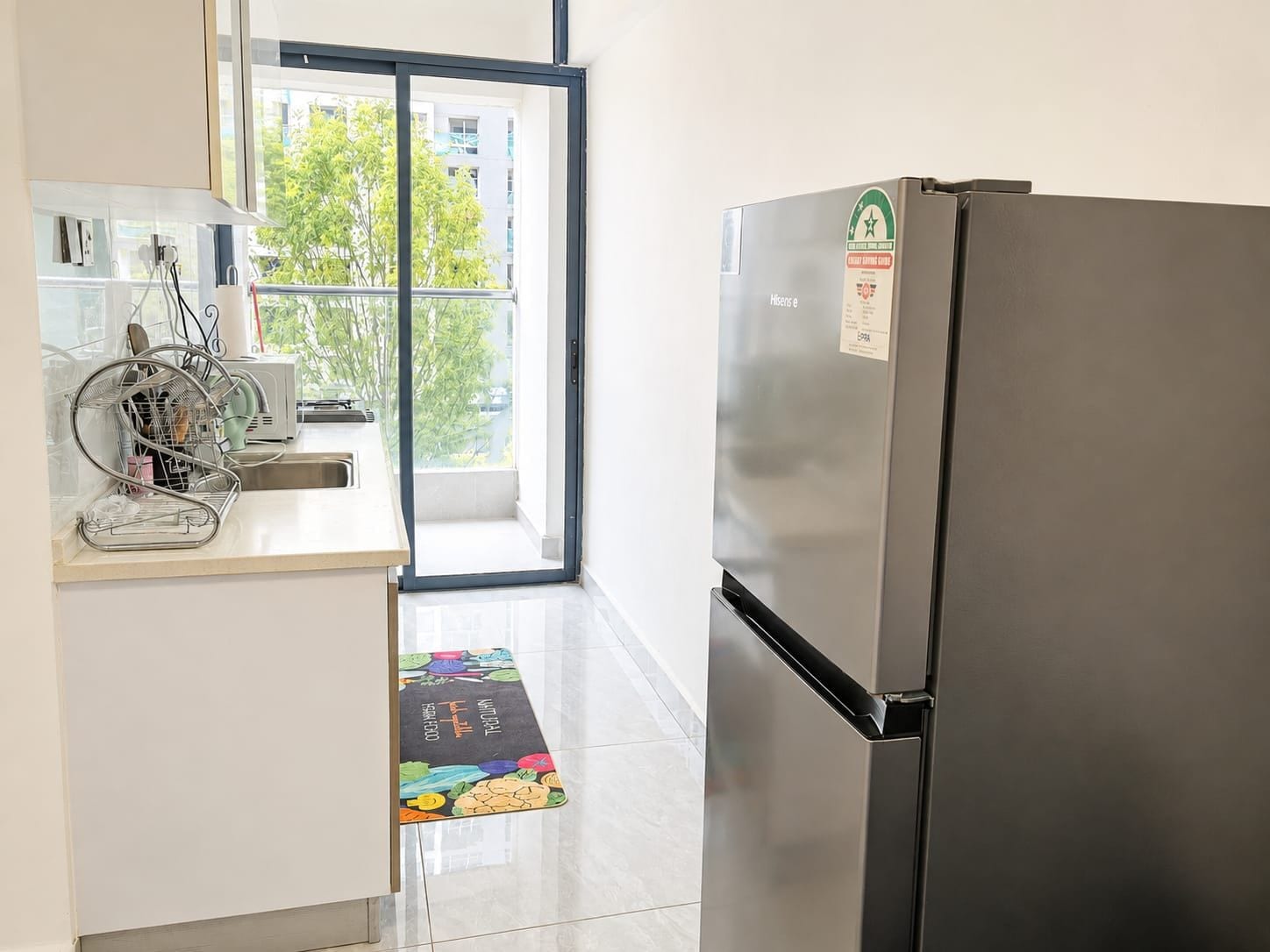 A kitchen with a stainless steel refrigerator, a dish rack, and a window with a view of buildings and trees.