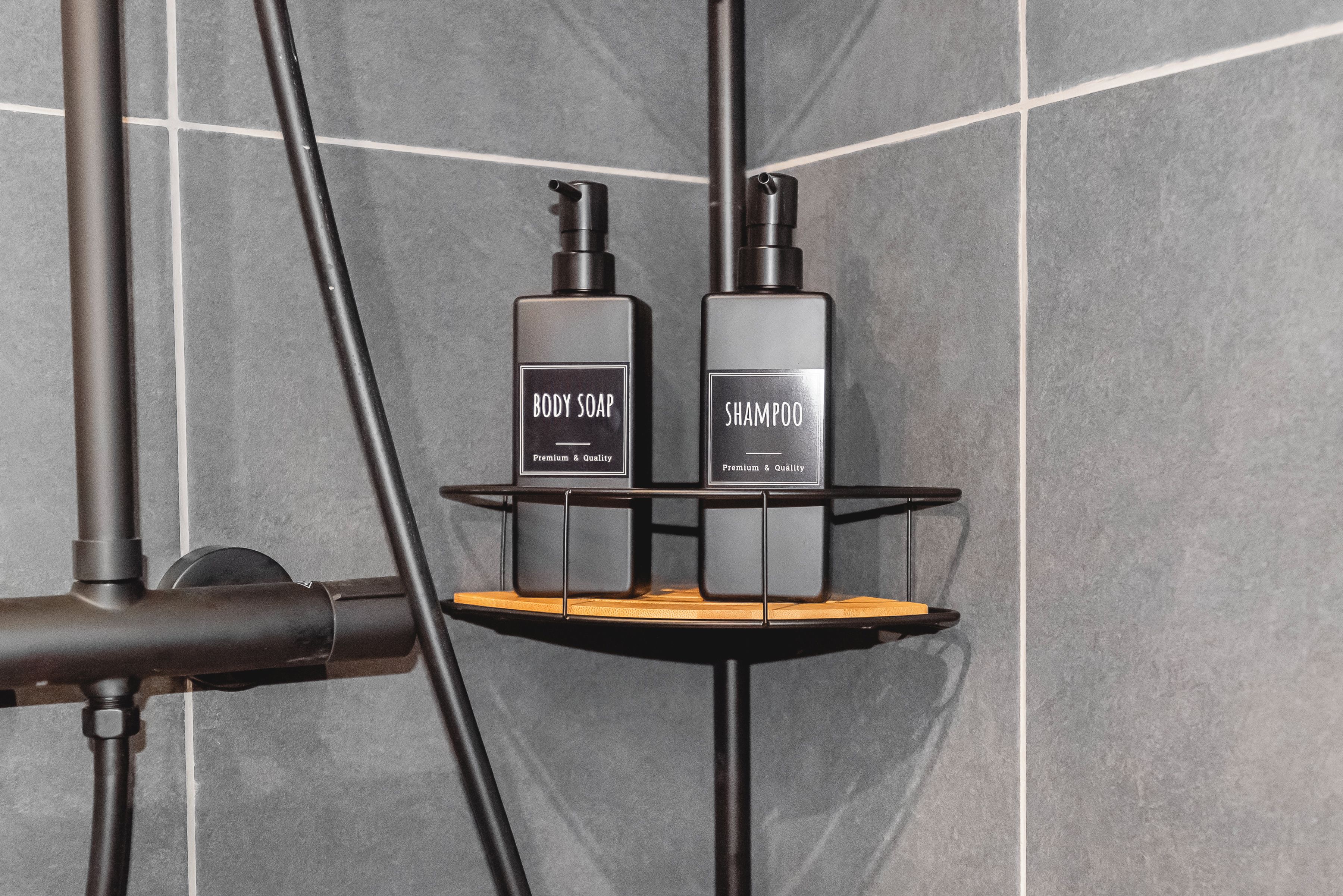 A bathroom with a shower head, a black towel rack, and two bottles of body soap and shampoo.