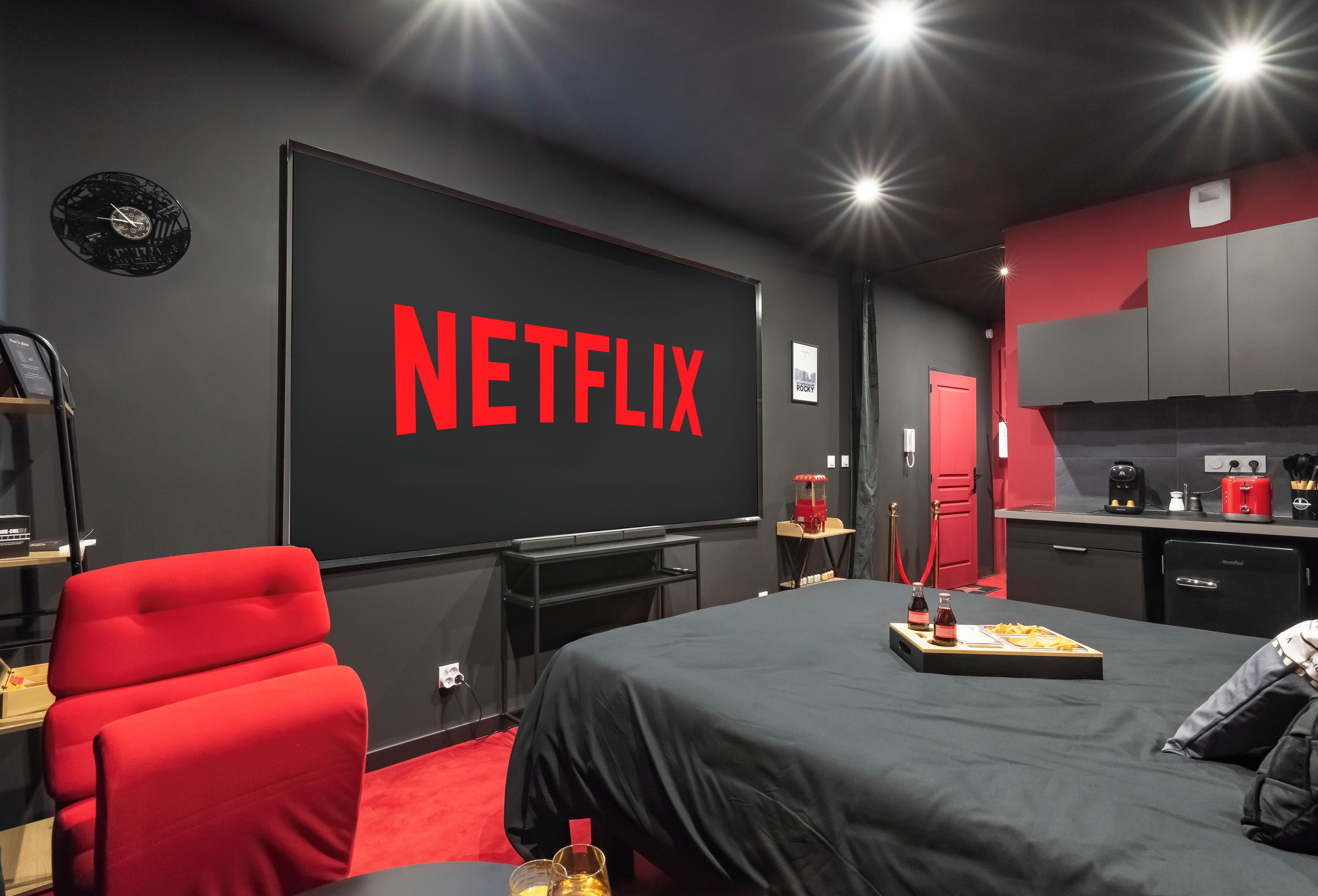 Una habitación moderna con un televisor de pantalla plana grande mostrando el logotipo de Netflix, una silla roja, una cama con colcha negra y una zona de kitchenette.
